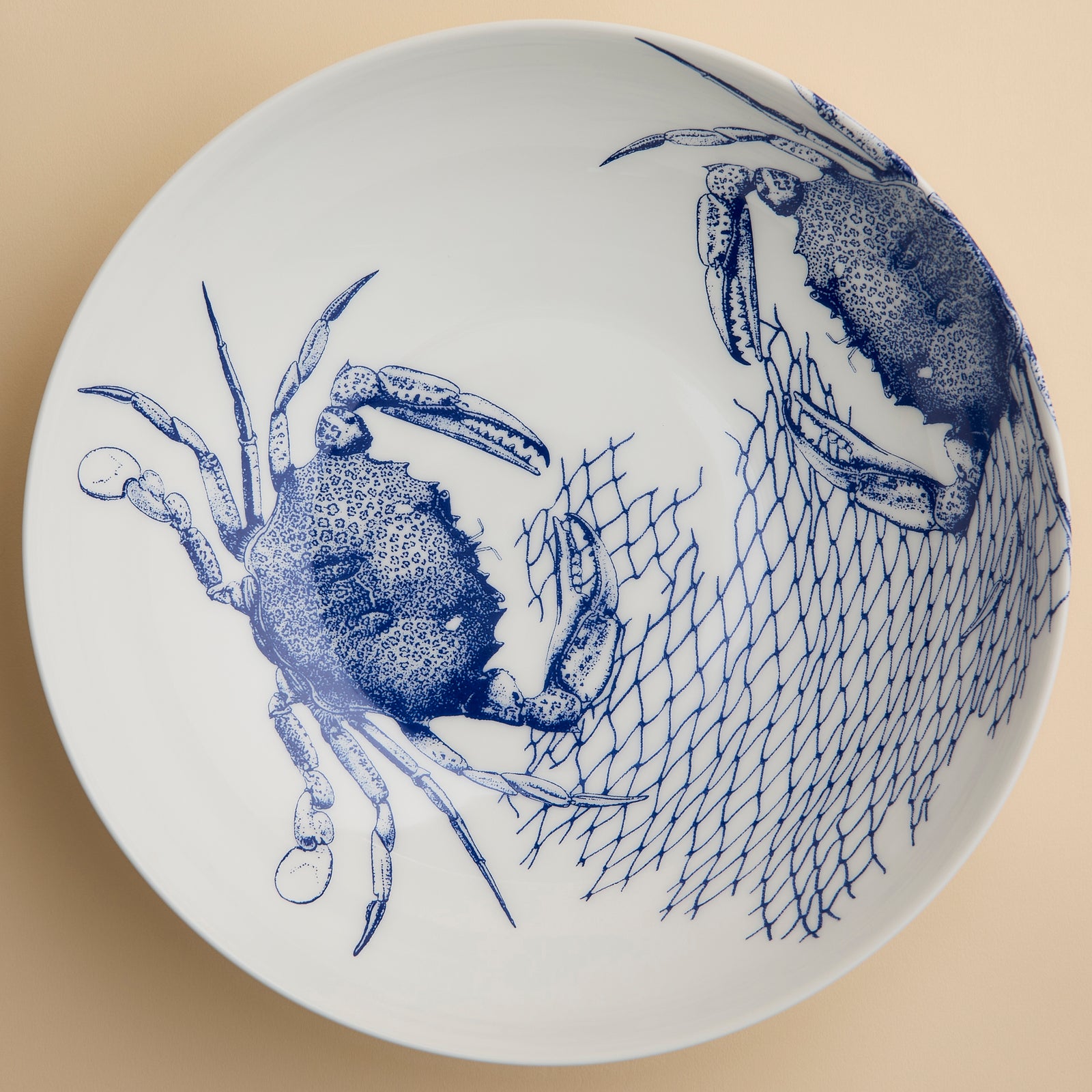 The Crab Wide Serving Bowl by Caskata Artisanal Home features white ceramic adorned with blue illustrations of two crabs and a fishing net on the right side, set against a beige background. Reminiscent of coastal charm, this piece is perfect as a decorative serving bowl for any seaside-inspired table setting.