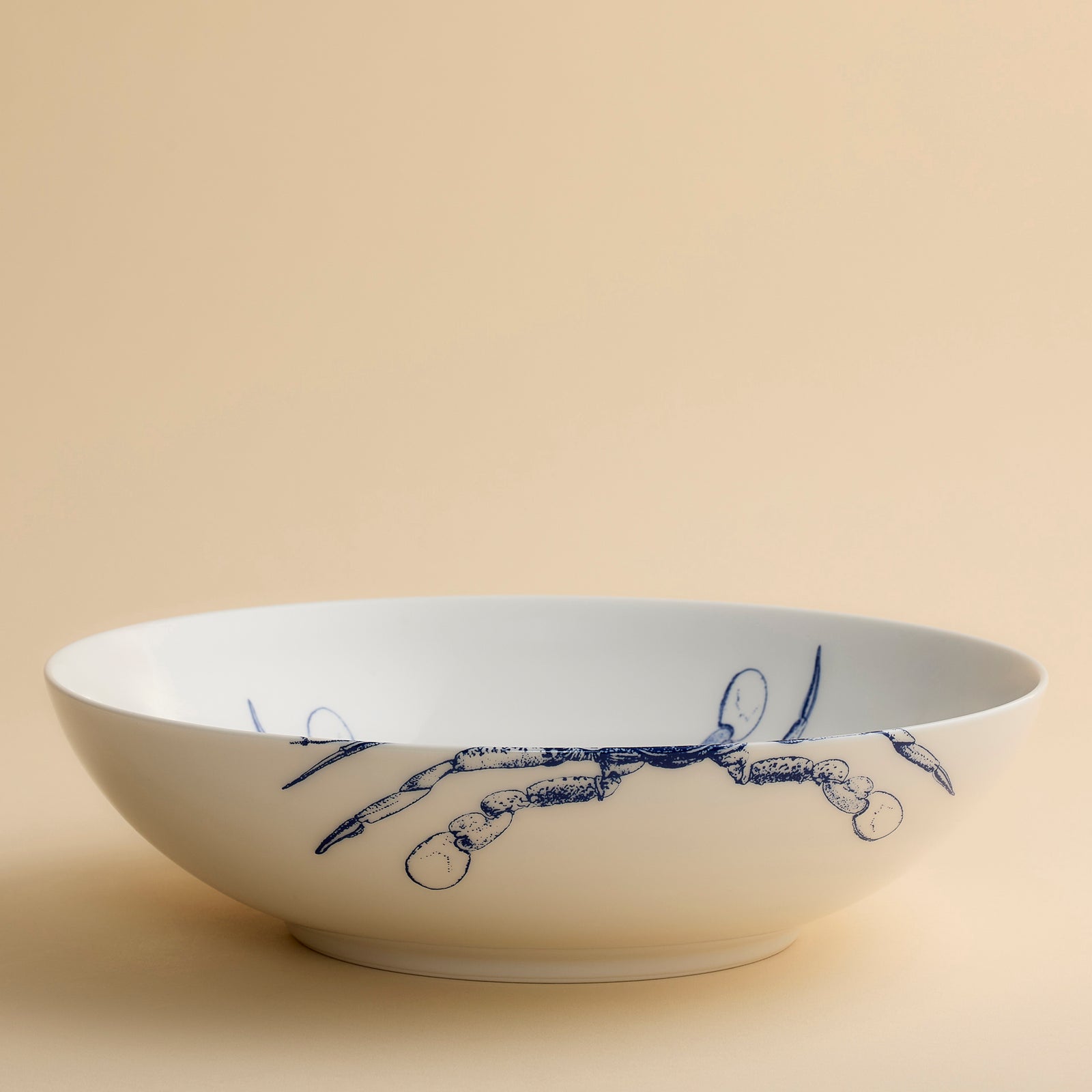 The Crab Wide Serving Bowl by Caskata Artisanal Home features white ceramic adorned with blue illustrations of two crabs and a fishing net on the right side, set against a beige background. Reminiscent of coastal charm, this piece is perfect as a decorative serving bowl for any seaside-inspired table setting.