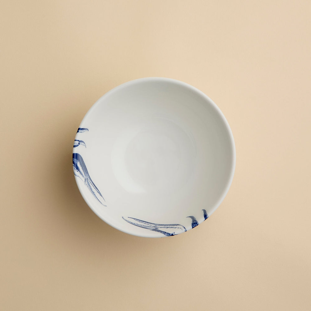 The Caskata Crab Cereal Bowl is a white ceramic bowl featuring a minimal blue crab claw design on the rim, displayed on a plain beige background.