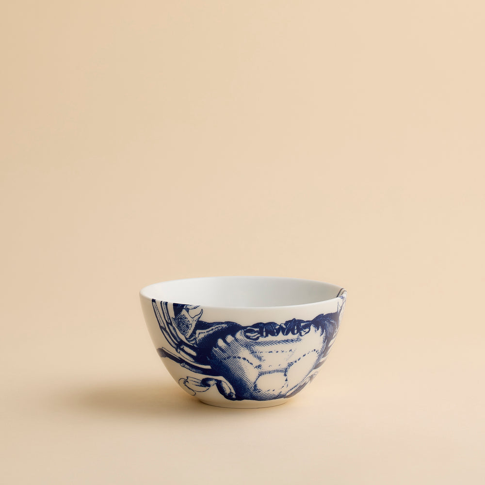 The Caskata Crab Cereal Bowl is a white ceramic bowl featuring a blue illustration of a crab on one side, displayed against a plain beige background.