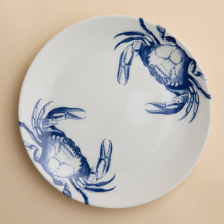 The Crab Coupe Dinner Plate by Caskata Artisanal Home showcases two blue crab illustrations on opposite sides of a white ceramic plate, set against a light beige background—an ideal choice for beach-inspired dining or bringing coastal charm to your table.