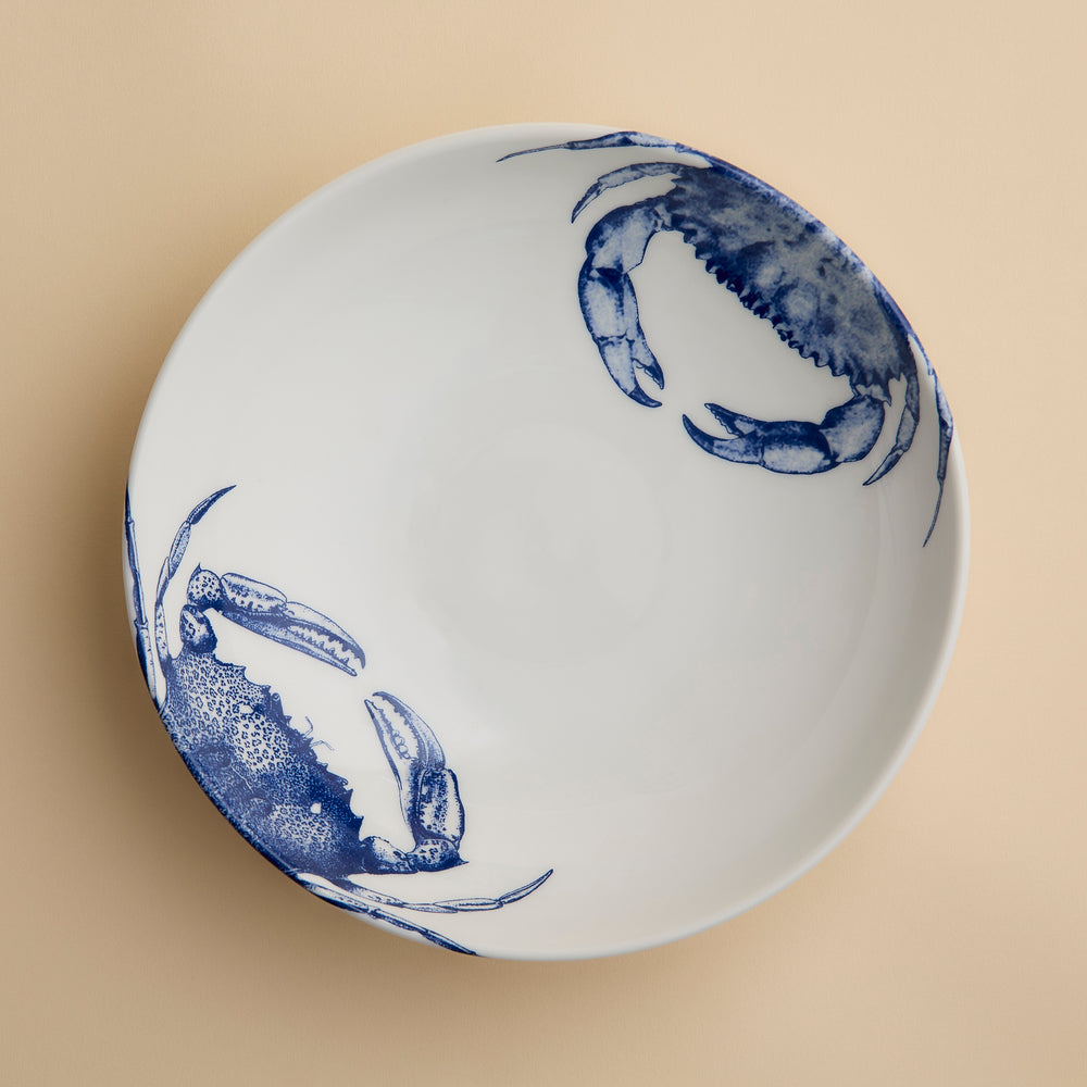 The Caskata Artisanal Home Crab Entrée Bowl is a white ceramic bowl featuring two blue crab illustrations on the rim and is pictured on a beige surface; this porcelain bowl is dishwasher safe for easy cleaning.