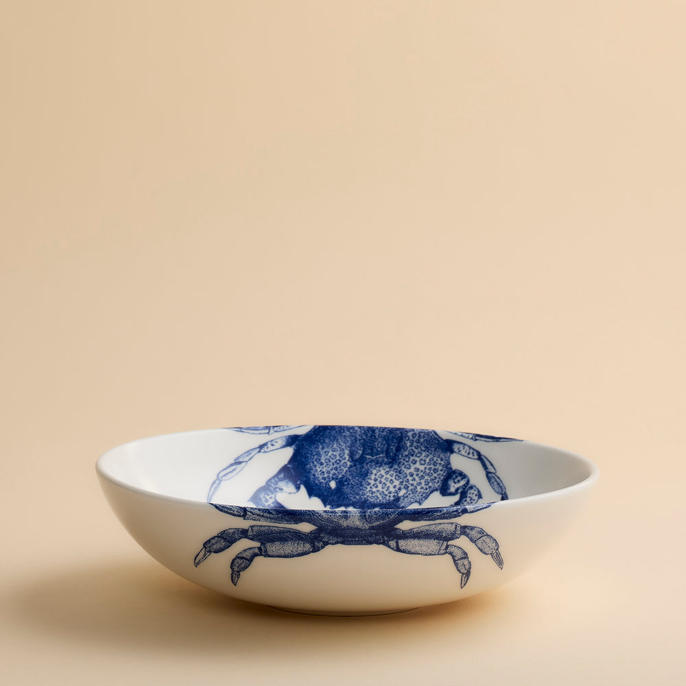 The Crab Entrée Bowl by Caskata Artisanal Home is a white porcelain bowl adorned with a blue crab illustration on the inside. Set on a beige surface against a plain beige background, this elegant bowl is dishwasher safe, making it ideal for everyday use.