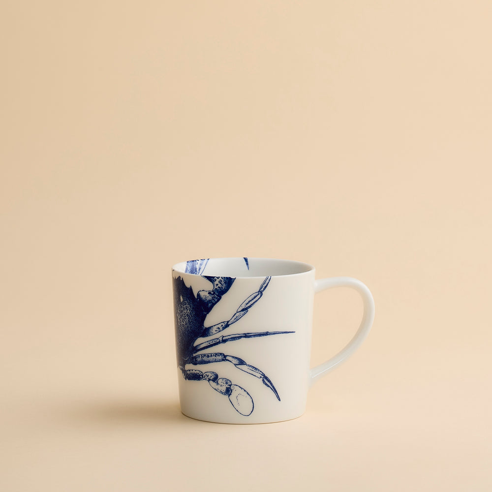 Discover the Caskata Artisanal Home Crab Mug—a white ceramic mug showcasing a charming blue crab illustration set against a simple beige background. Ideal for anyone who appreciates unique crab mug designs.