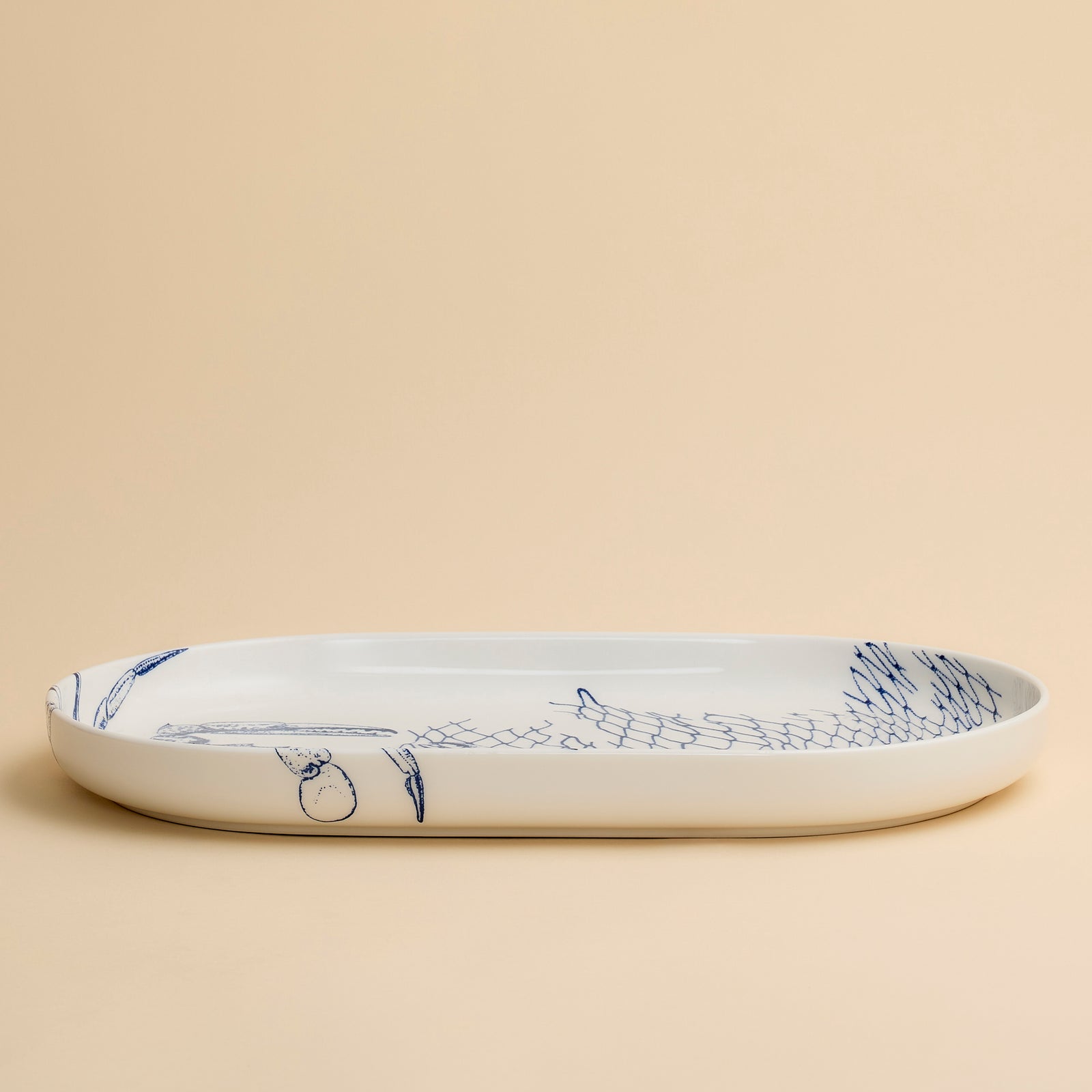 The Caskata Crab Large Coupe Oval Platter showcases a striking blue crab and fishing net illustration on a beige porcelain background, bringing coastal elegance to any table setting.