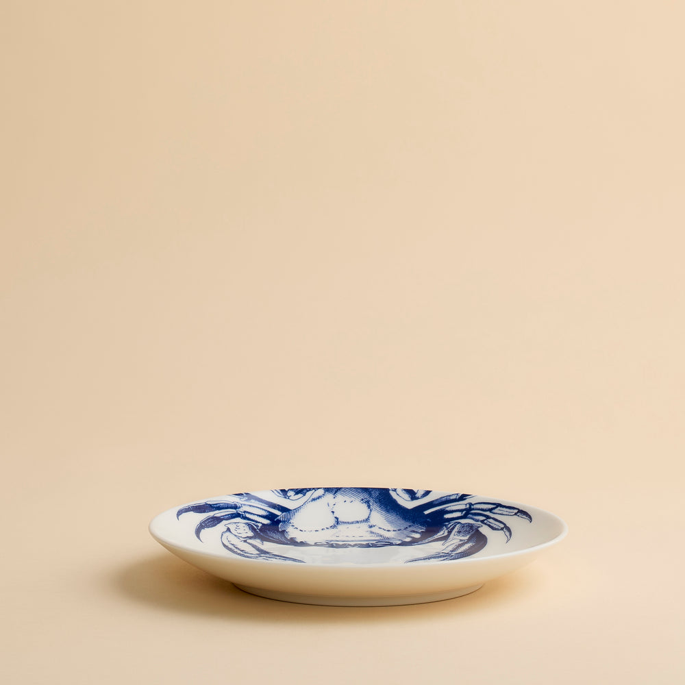 Caskata Artisanal Home’s Crab Coupe Salad Plate features a charming blue crab illustration on white ceramic, set against a beige background—an ideal addition to your coastal dinnerware collection.