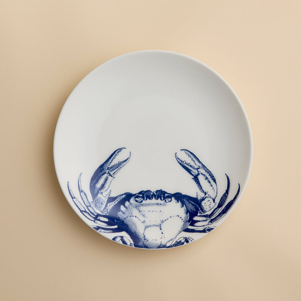 The Crab Coupe Salad Plate by Caskata Artisanal Home features a white porcelain design accented with a blue crab illustration on the lower half, making it a perfect addition to your coastal dinnerware collection for serving fresh salads or appetizers.