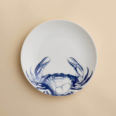 The Crab Coupe Salad Plate by Caskata Artisanal Home features a white porcelain design accented with a blue crab illustration on the lower half, making it a perfect addition to your coastal dinnerware collection for serving fresh salads or appetizers.