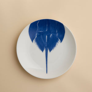 The Horseshoe Crab Coupe Salad Plate by Caskata Artisanal Home features a blue illustration of a horseshoe crab, seen from above, on a modern white coupe plate with its tail extending toward the bottom edge.