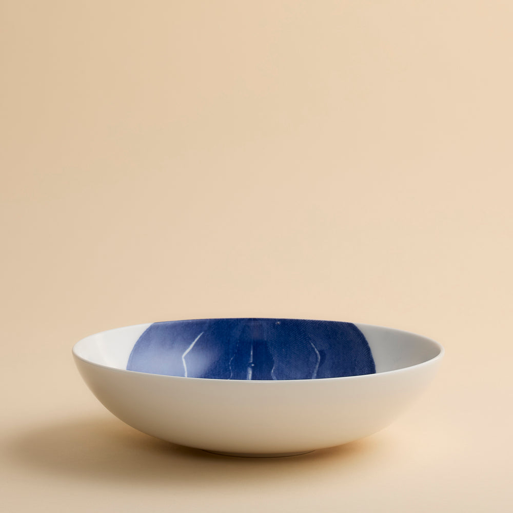 A white ceramic Caskata Horseshoe Crab Entrée Bowl, featuring a wide and shallow shape with a dark blue painted interior design, displayed against a plain beige background.