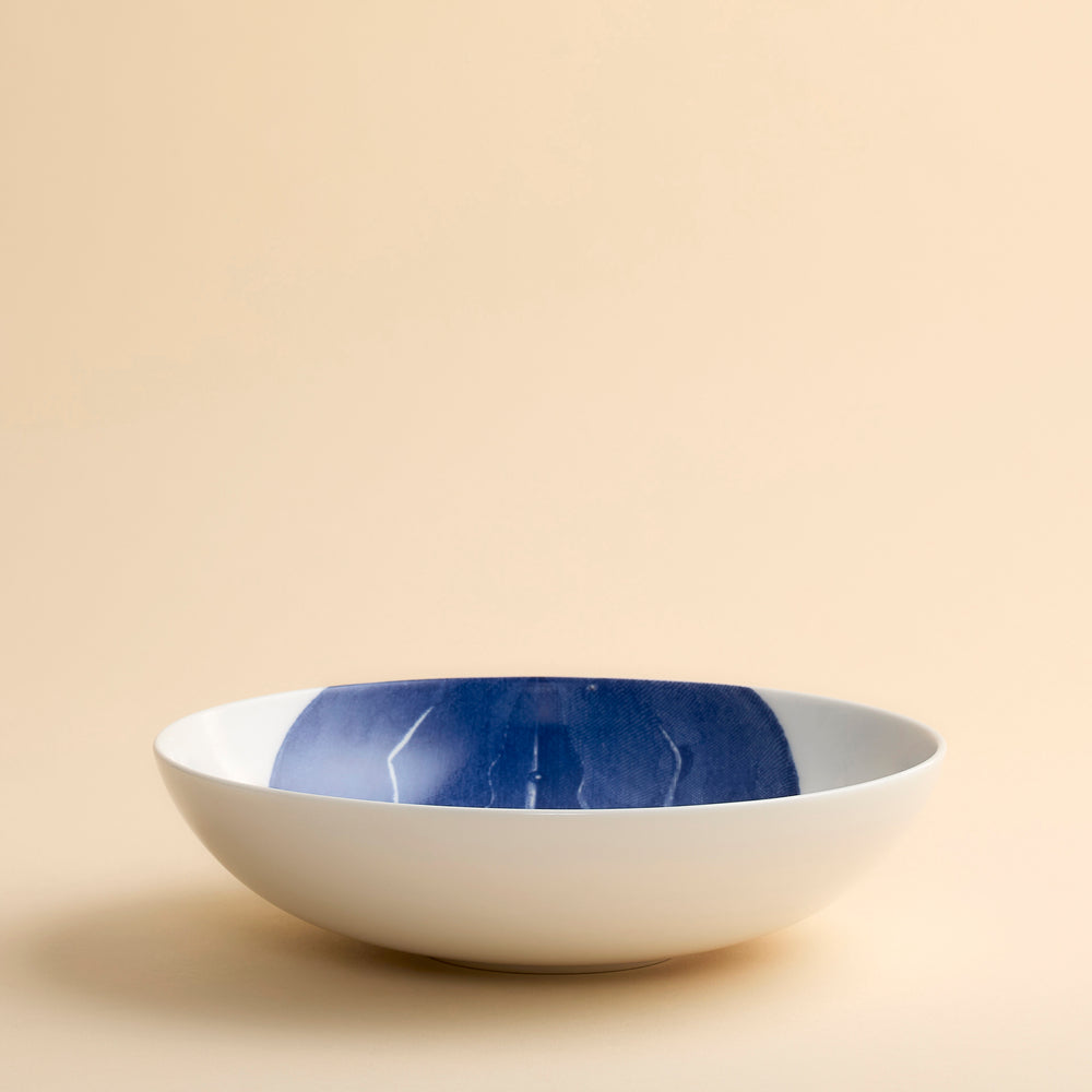A white porcelain Caskata Horseshoe Crab Entrée Bowl, featuring a wide and shallow shape with a dark blue detailed engraving of a horseshoe design, displayed against a plain beige background.