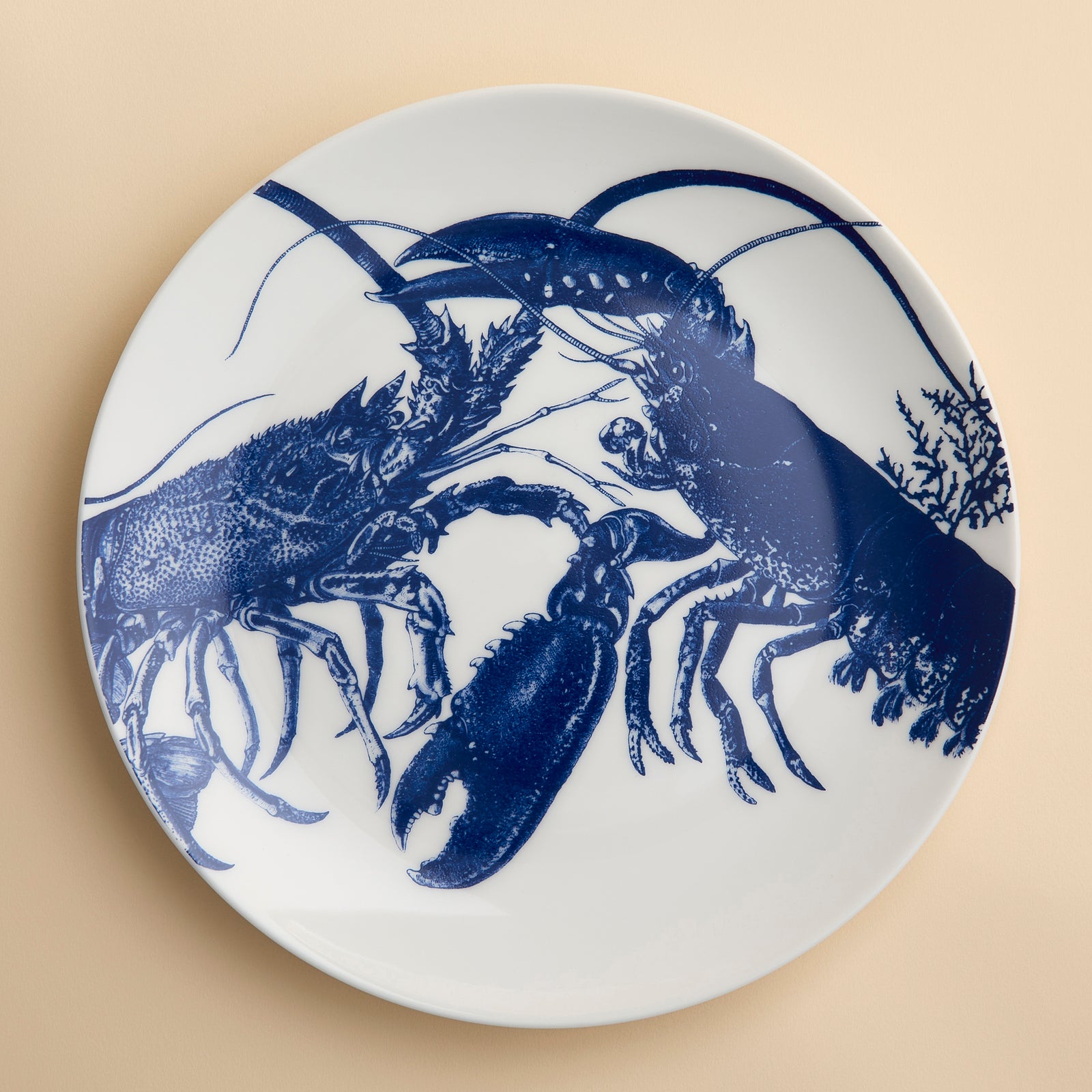 Blue lobsters, one from the East, one from the West, lock in a graphic embrace across the face of this coupe porcelain dinner plate from Caskata.