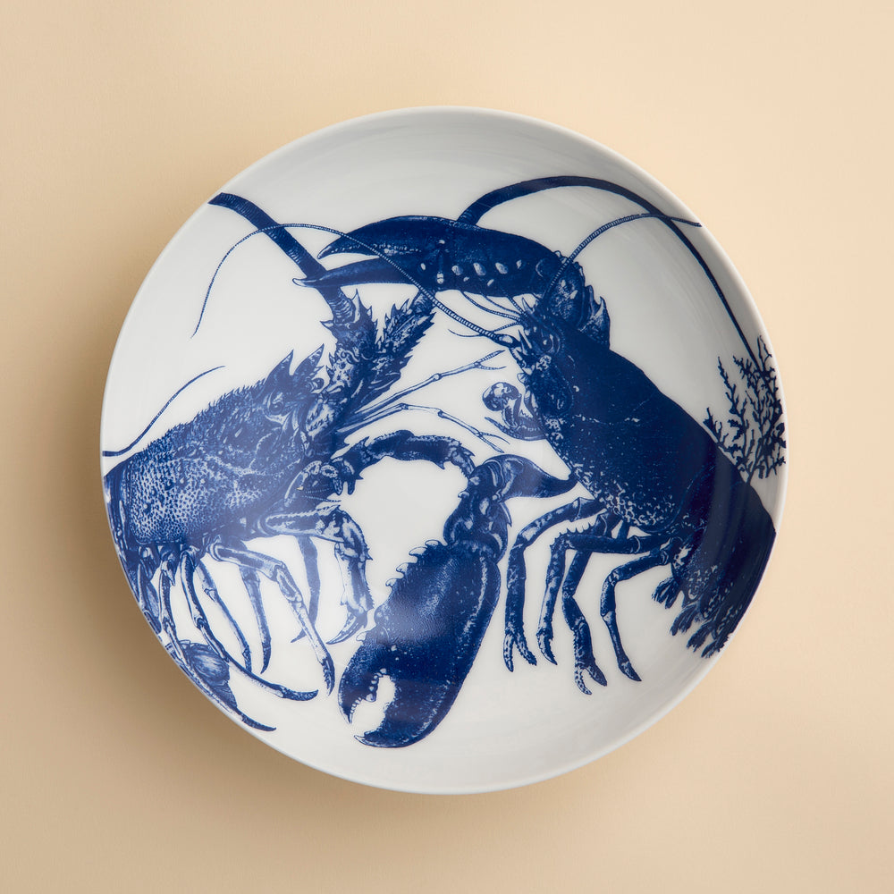 The Lobster Entrée Bowl by Caskata, from our coastal dinnerware collection, is a white ceramic bowl featuring a blue illustration of two lobsters facing each other on a beige background.