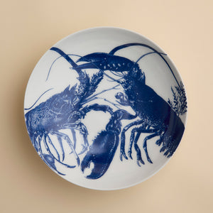 The Lobster Entrée Bowl by Caskata, from our coastal dinnerware collection, is a white ceramic bowl featuring a blue illustration of two lobsters facing each other on a beige background.