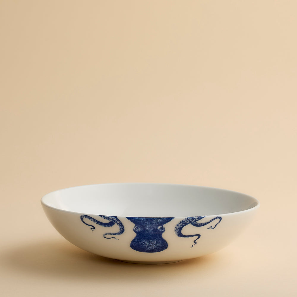 The Lucy Entrée Bowl by Caskata Artisanal Home is a white porcelain bowl featuring blue painted designs, including two dragons and a central vase motif, set against a plain beige background.
