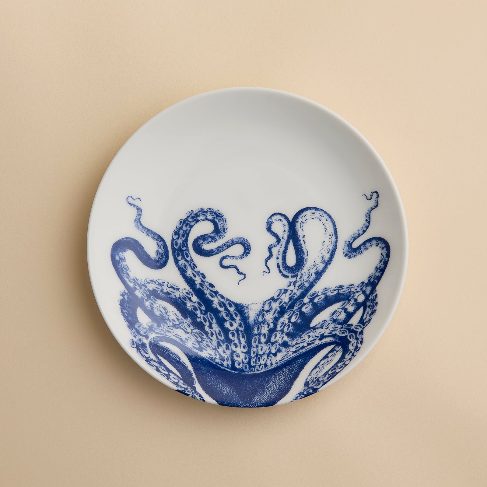 The Lucy Coupe Salad Plate by Caskata Artisanal Home is a white porcelain coupe plate adorned with blue octopus tentacles extending outward on a beige background; this unique salad plate combines stylish design with dishwasher-safe convenience.
