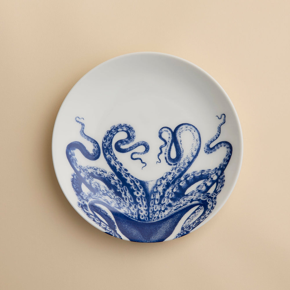 The Lucy Coupe Salad Plate by Caskata Artisanal Home is a white porcelain coupe plate adorned with blue octopus tentacles extending outward on a beige background; this unique salad plate combines stylish design with dishwasher-safe convenience.
