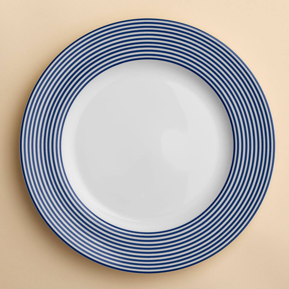 A blue and white Newport Stripe Rimmed Dinner Plate by Caskata Artisanal Home with concentric blue circles around the edge, perfect for contemporary porcelain dinnerware collections shown against a creamy beige background.
