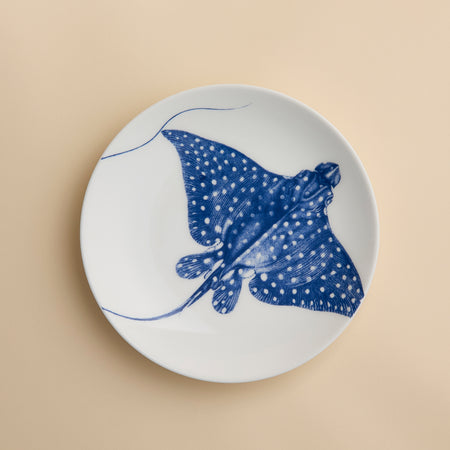 The Caskata Stingray Coupe Salad Plate, crafted from white porcelain and featuring a striking blue illustration of a spotted stingray at its center, infuses coastal charm and elegant style into your table setting. Shown on a light beige surface, this plate beautifully merges seaside inspiration with refined design.