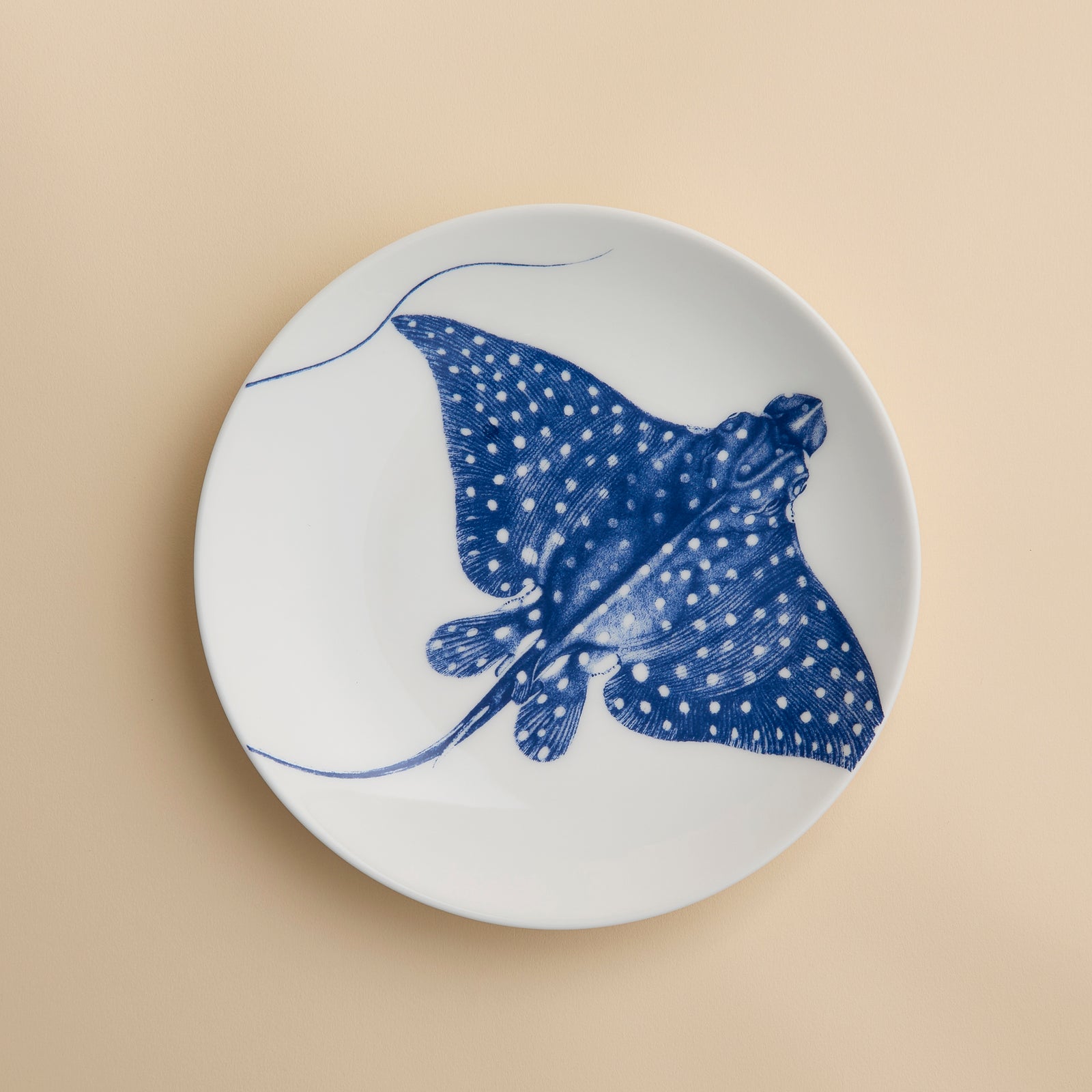 The Caskata Stingray Coupe Salad Plate, crafted from white porcelain and featuring a striking blue illustration of a spotted stingray at its center, infuses coastal charm and elegant style into your table setting. Shown on a light beige surface, this plate beautifully merges seaside inspiration with refined design.