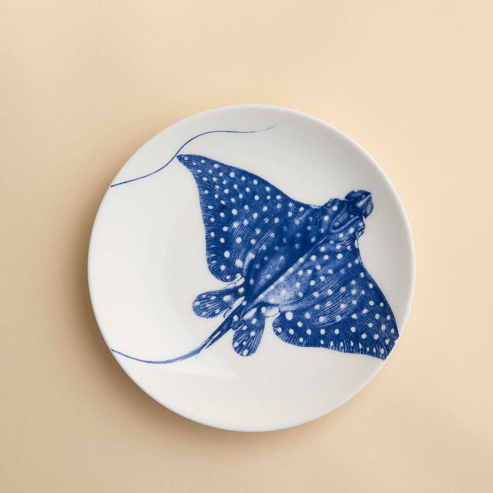 The Caskata Stingray Coupe Salad Plate, crafted from white porcelain and featuring a striking blue illustration of a spotted stingray at its center, infuses coastal charm and elegant style into your table setting. Shown on a light beige surface, this plate beautifully merges seaside inspiration with refined design.