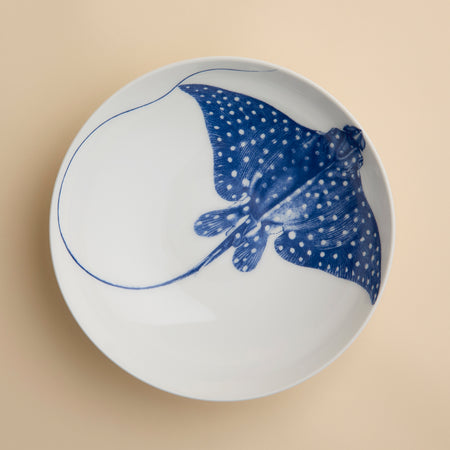 The Ray the Stingray Entrée Bowl by Caskata is a white ceramic bowl, ideal for stingray-themed dinnerware collections, and showcases a blue illustration of a spotted stingray viewed from above, elegantly positioned on a beige surface.