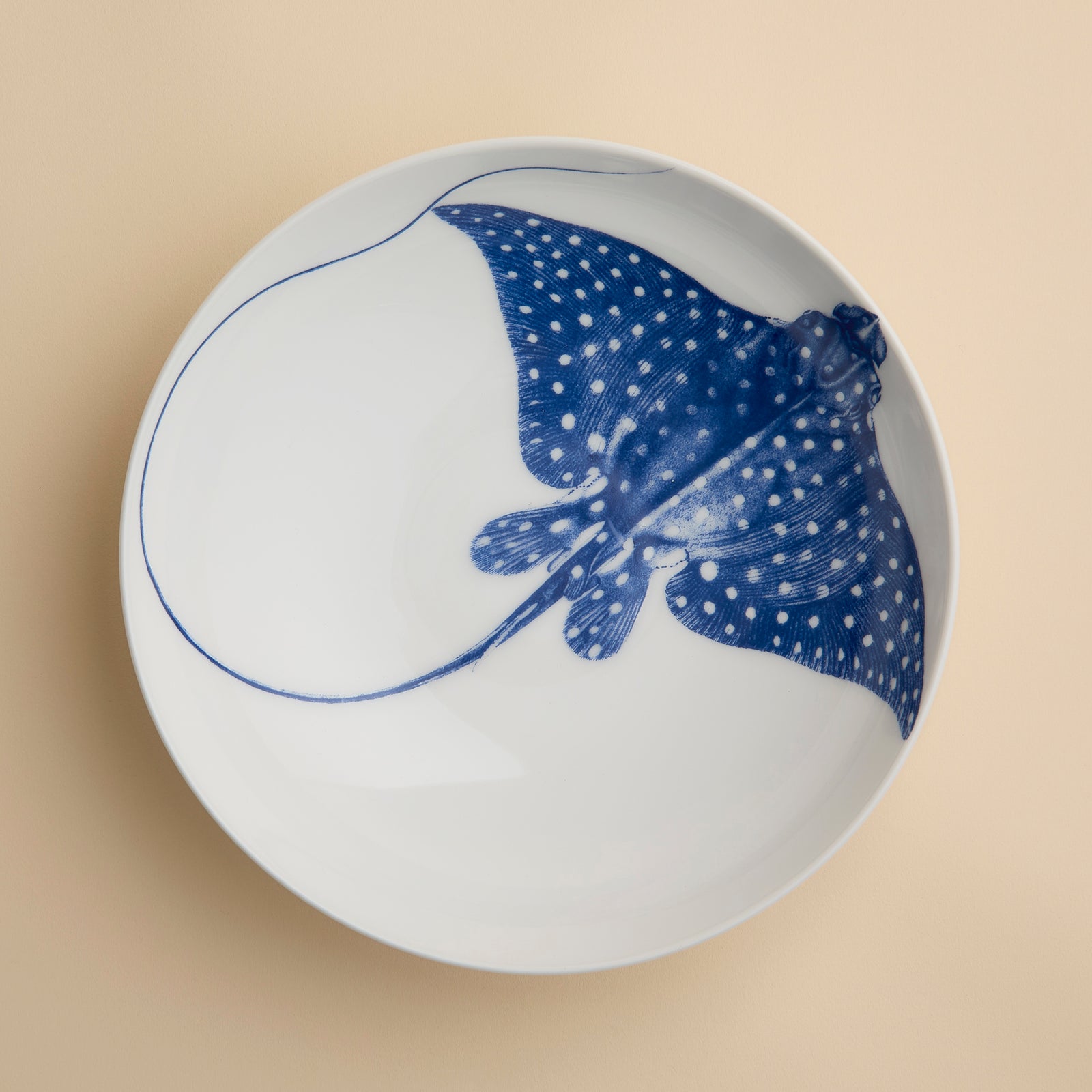 The Ray the Stingray Entrée Bowl by Caskata is a white ceramic bowl, ideal for stingray-themed dinnerware collections, and showcases a blue illustration of a spotted stingray viewed from above, elegantly positioned on a beige surface.