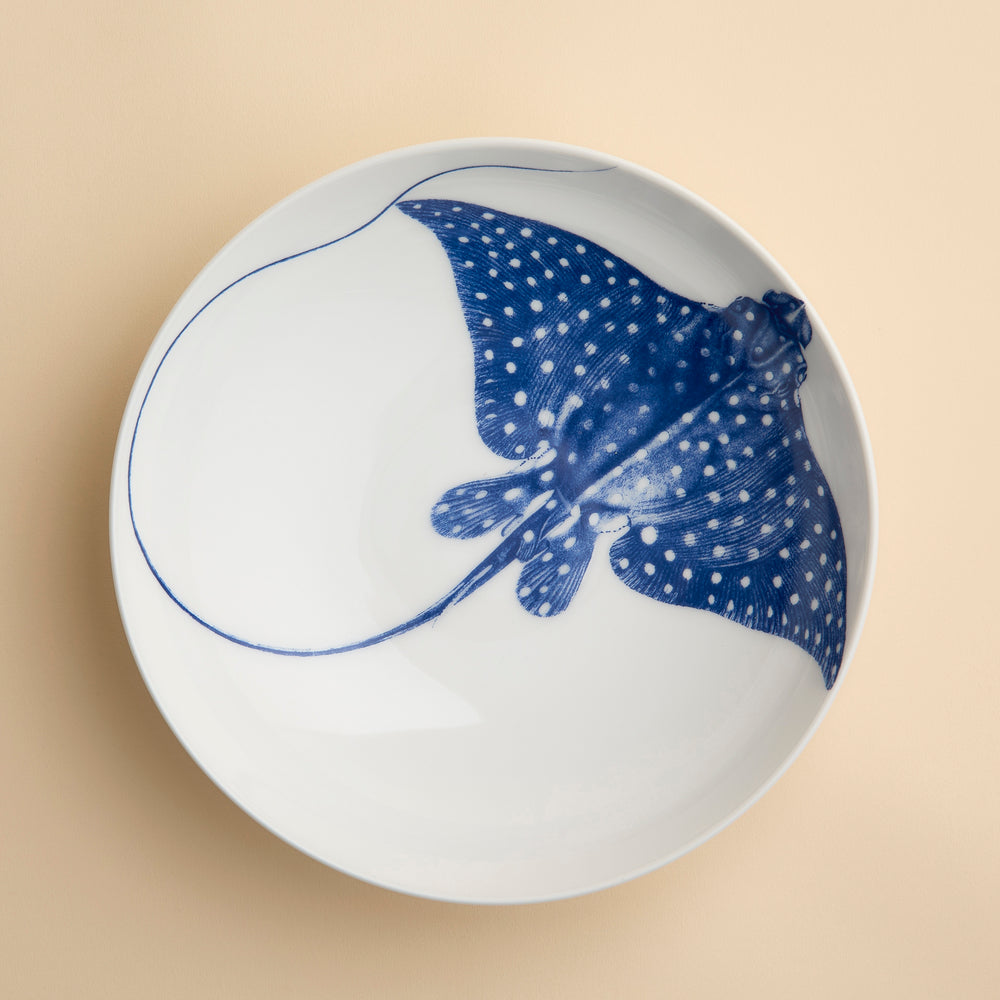 The Ray the Stingray Entrée Bowl by Caskata is a white ceramic bowl, ideal for stingray-themed dinnerware collections, and showcases a blue illustration of a spotted stingray viewed from above, elegantly positioned on a beige surface.