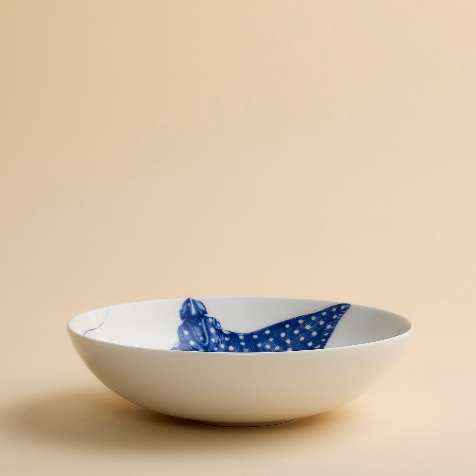 The Ray the Stingray Entrée Bowl by Caskata is a white ceramic bowl, ideal for stingray-themed dinnerware collections, and showcases a blue illustration of a spotted stingray viewed from above, elegantly positioned on a beige surface.