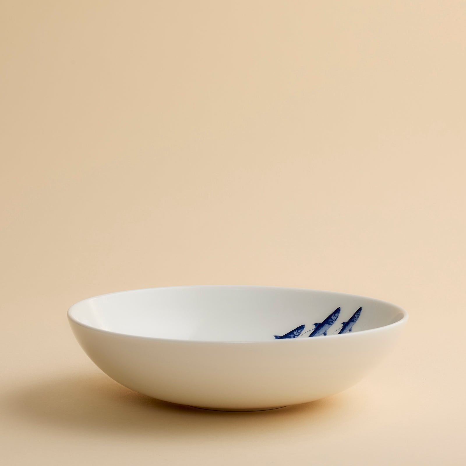 The School Fish Entrée Bowl by Caskata Artisanal Home features a white ceramic design adorned with a group of blue fish, seen from above on a beige background—an ideal coastal porcelain piece for serving your favorite seafood or showcasing as elegant blue fish dishware.