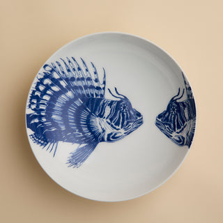 The Spiky Fish Entrée Bowl by Caskata features two blue fish illustrations facing each other on a white ceramic base, set against a plain beige background. Perfect for lovers of coastal dinnerware, this bowl adds a hint of maritime charm to any table setting.