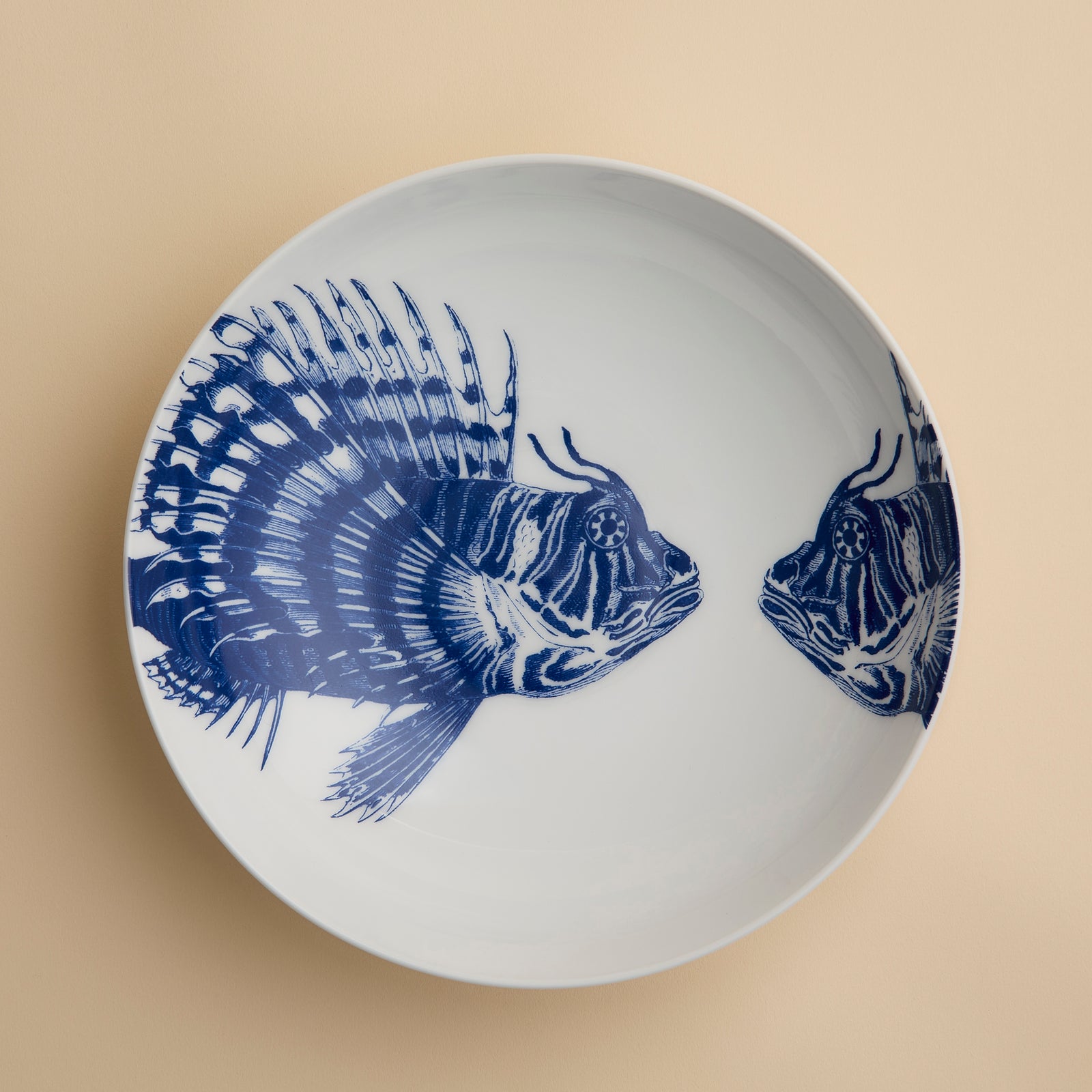 The Spiky Fish Entrée Bowl by Caskata features two blue fish illustrations facing each other on a white ceramic base, set against a plain beige background. Perfect for lovers of coastal dinnerware, this bowl adds a hint of maritime charm to any table setting.