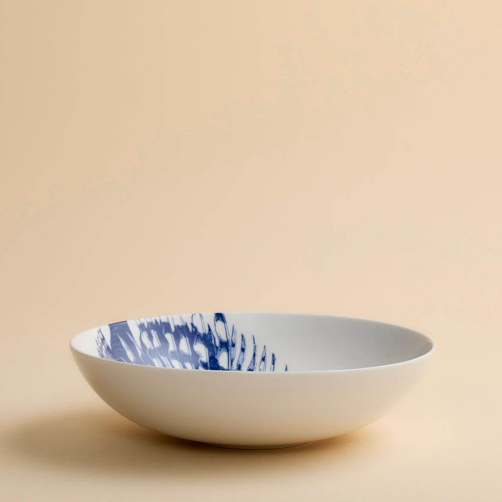 The Caskata Spiky Fish Entrée Bowl is a shallow white porcelain bowl adorned with a striking blue abstract fish motif on one side, capturing the coastal charm of elegant dinnerware and shown here against a simple beige background.