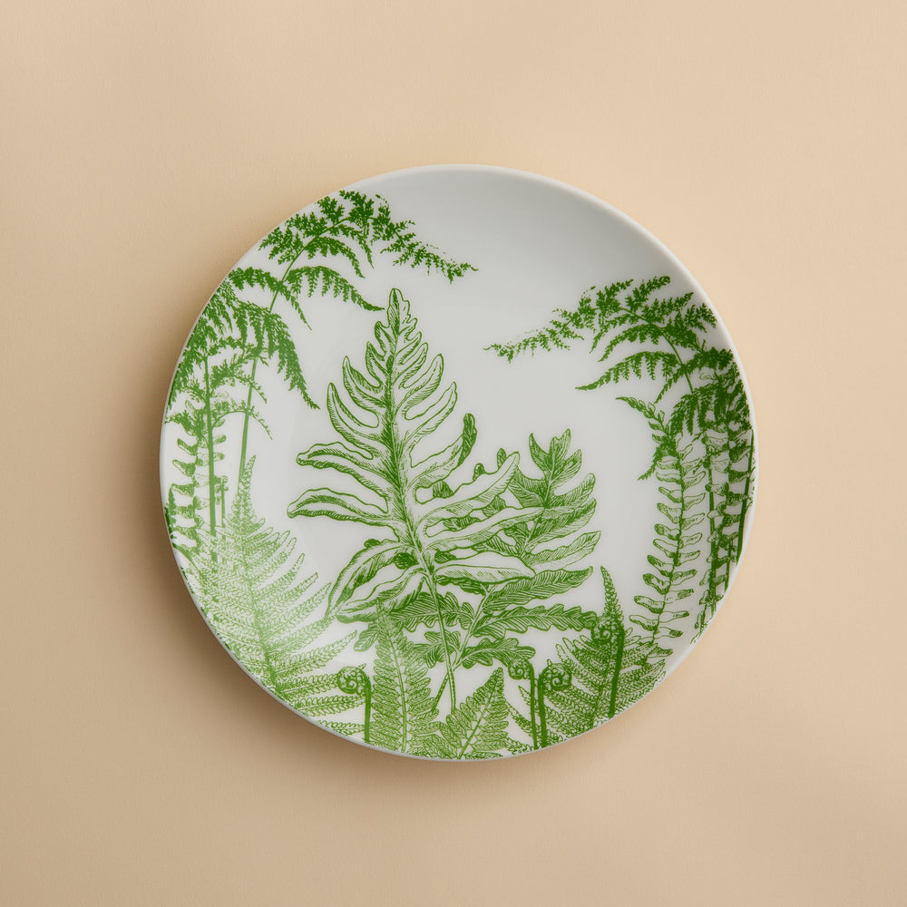 A round white plate adorned with green fern leaf illustrations rests against a beige background—Caskata’s Spring Verde Coupe Salad Plate showcases a charming botanical motif that lends elegance to any table setting. Expertly crafted from premium porcelain, this salad plate blends style with lasting durability.