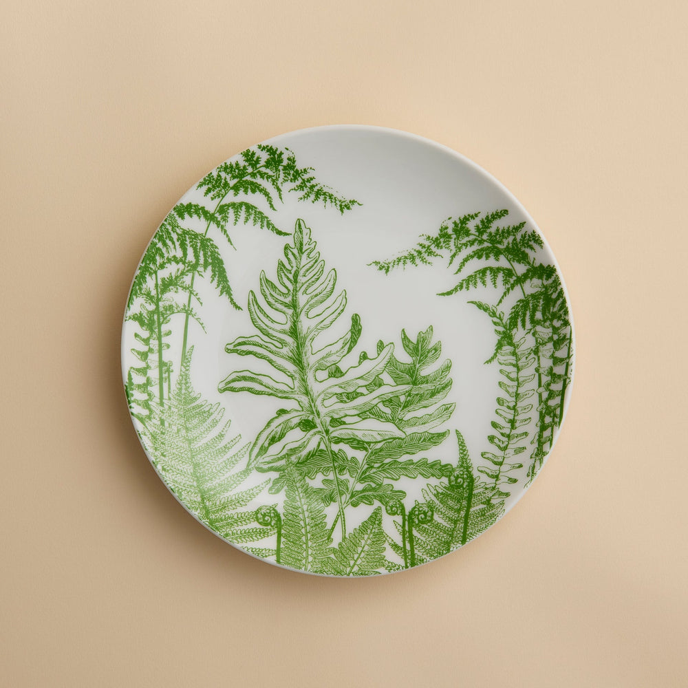 A round white plate adorned with green fern leaf illustrations rests against a beige background—Caskata’s Spring Verde Coupe Salad Plate showcases a charming botanical motif that lends elegance to any table setting. Expertly crafted from premium porcelain, this salad plate blends style with lasting durability.
