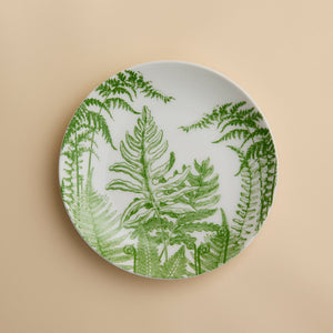 A round white plate adorned with green fern leaf illustrations rests against a beige background—Caskata’s Spring Verde Coupe Salad Plate showcases a charming botanical motif that lends elegance to any table setting. Expertly crafted from premium porcelain, this salad plate blends style with lasting durability.