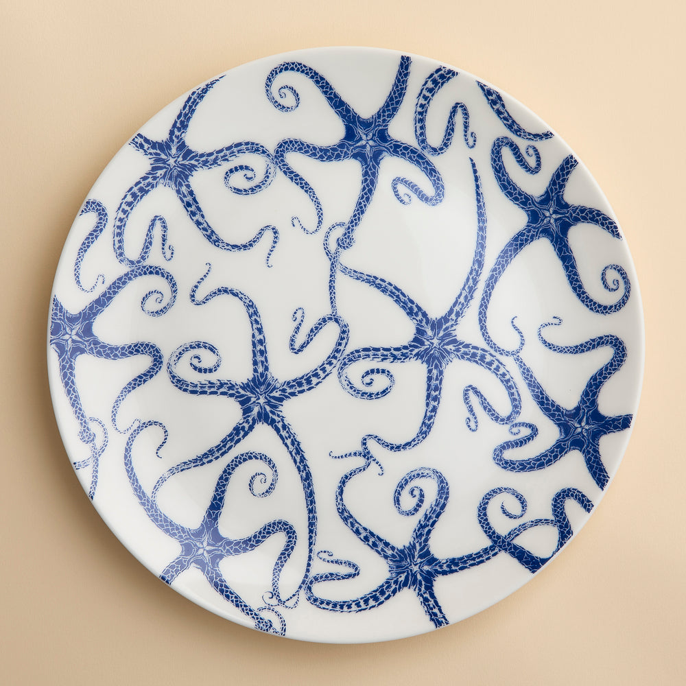 A Caskata Starfish Coupe Dinner Plate featuring a contemporary shape and a pattern of blue whimsical starfish dancing across the face in blue on creamy white porcelain shown on a beige background