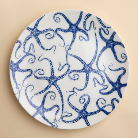 A Caskata Starfish Coupe Dinner Plate featuring a contemporary shape and a pattern of blue whimsical starfish dancing across the face in blue on creamy white porcelain shown on a beige background