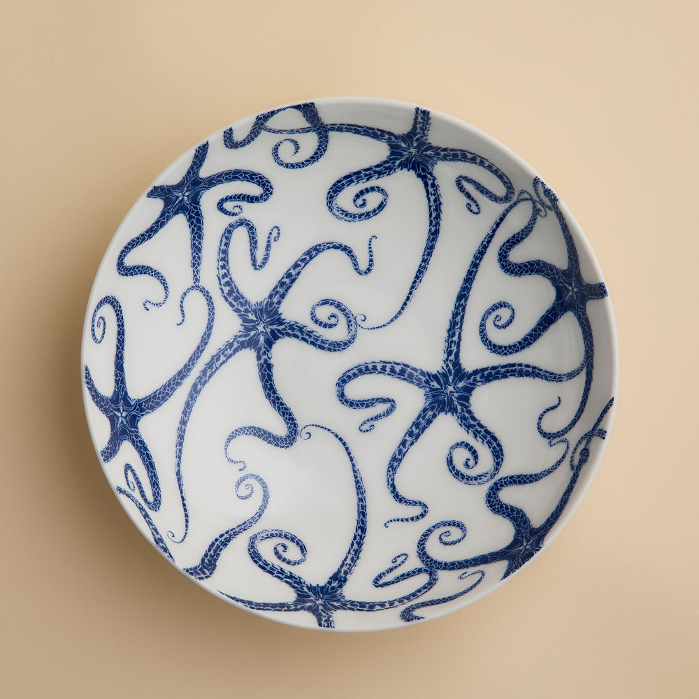Caskata’s Starfish Entrée Bowl, viewed from above against a light beige background, features a white ceramic design adorned with a blue starfish motif, capturing the essence of coastal charm.