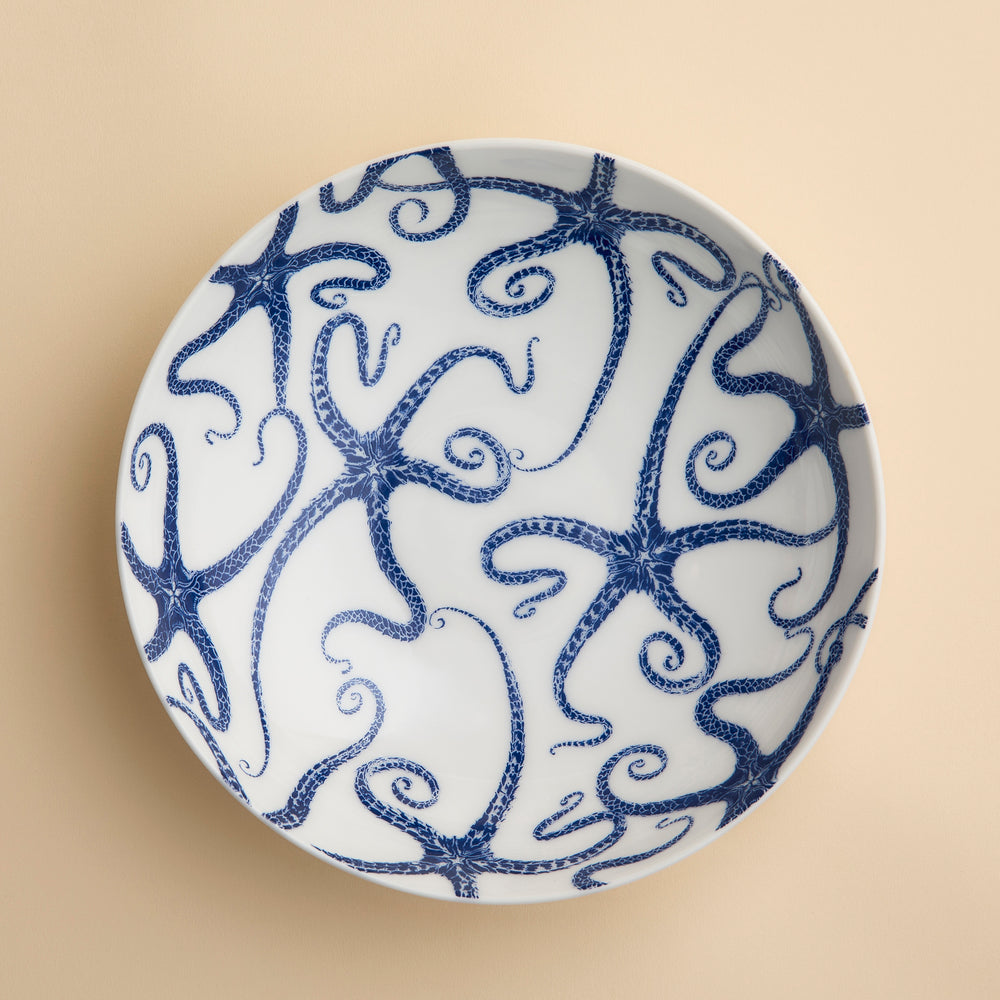 Caskata’s Starfish Entrée Bowl, viewed from above against a light beige background, features a white ceramic design adorned with a blue starfish motif, capturing the essence of coastal charm.