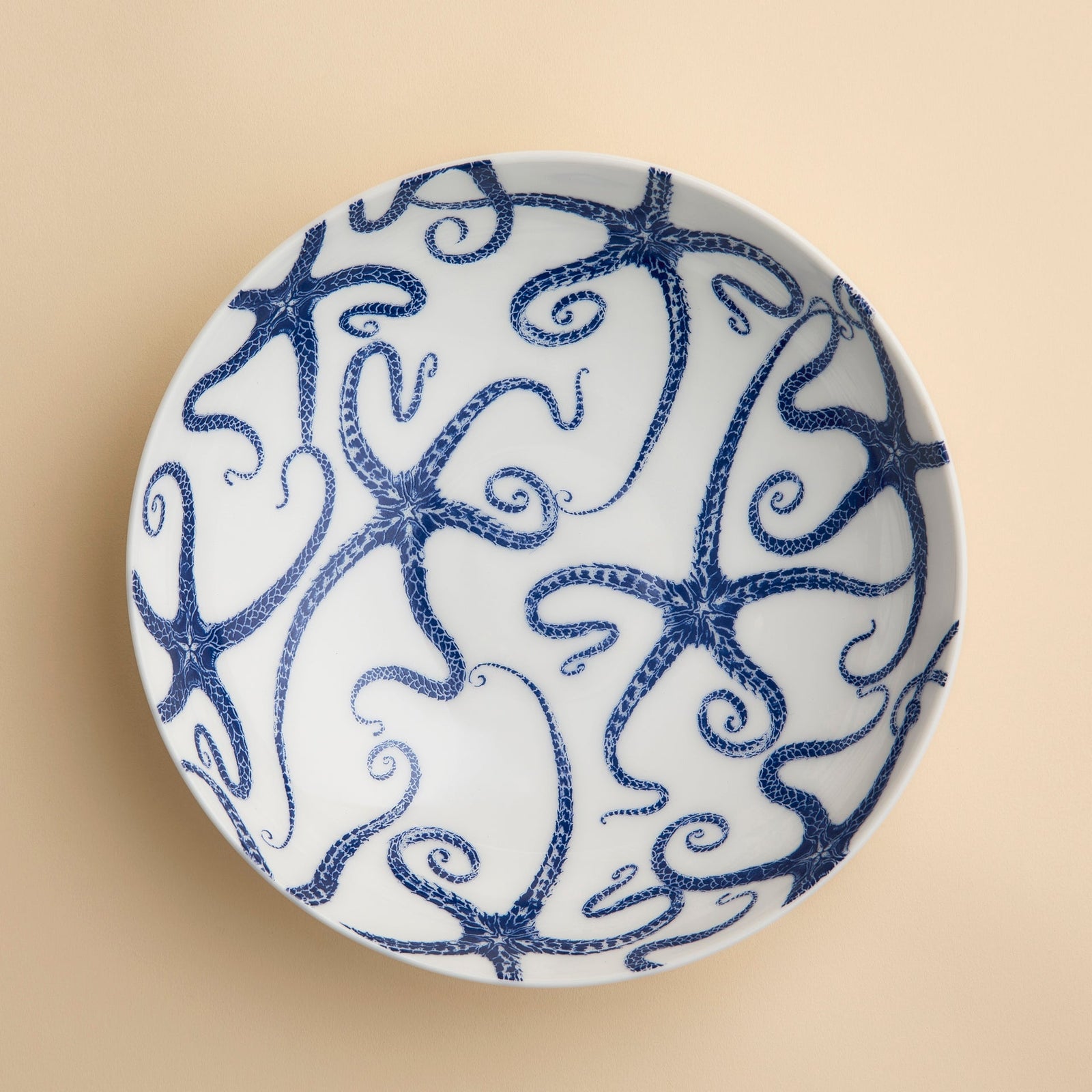 Caskata’s Starfish Entrée Bowl, viewed from above against a light beige background, features a white ceramic design adorned with a blue starfish motif, capturing the essence of coastal charm.