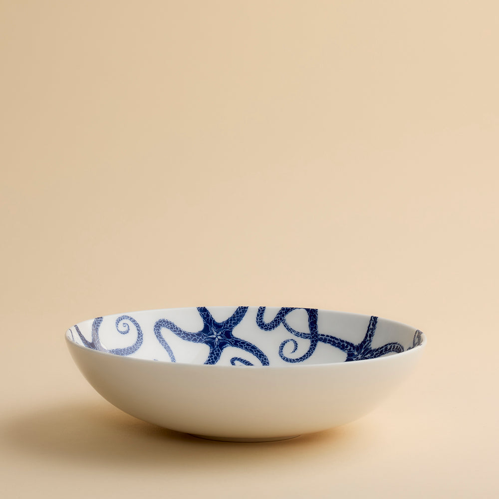 The Caskata Starfish Entrée Bowl is a shallow white porcelain serving bowl adorned with a striking blue starfish pattern on the inside, shown here on a neutral beige surface—a coastal-inspired piece that elevates your table setting.