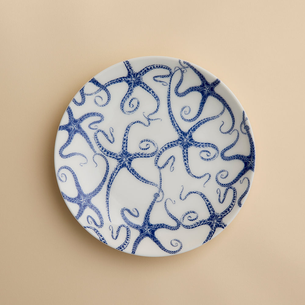 A round white porcelain salad plate featuring a blue starfish motif from Caskata Artisanal Home’s Starfish Coupe Salad Plate collection is displayed against a plain beige background, highlighting its ocean-inspired dinnerware design.