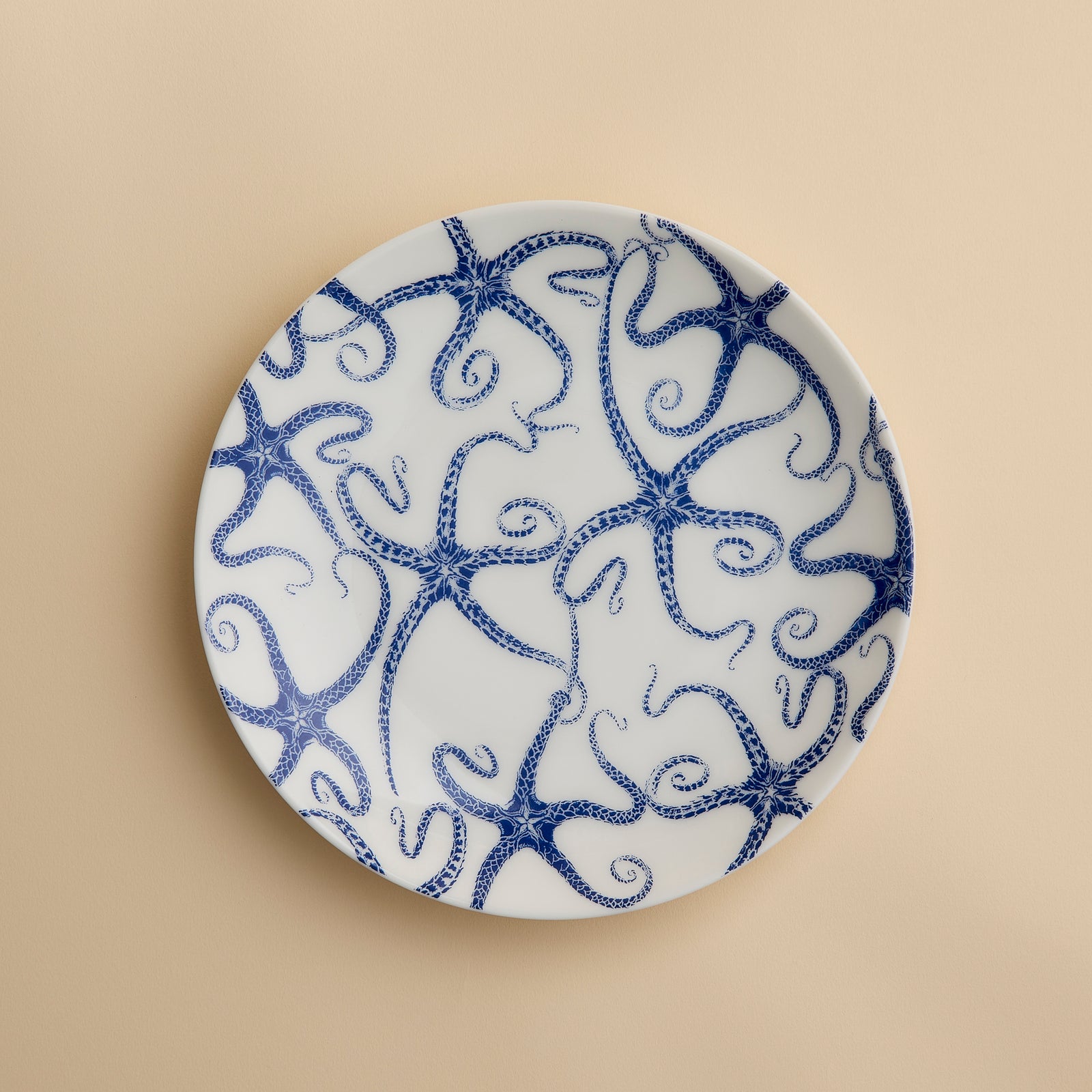 A round white porcelain salad plate featuring a blue starfish motif from Caskata Artisanal Home’s Starfish Coupe Salad Plate collection is displayed against a plain beige background, highlighting its ocean-inspired dinnerware design.