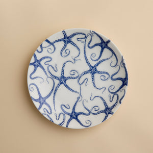 A round white porcelain salad plate featuring a blue starfish motif from Caskata Artisanal Home’s Starfish Coupe Salad Plate collection is displayed against a plain beige background, highlighting its ocean-inspired dinnerware design.