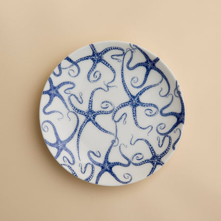 A round white porcelain salad plate featuring a blue starfish motif from Caskata Artisanal Home’s Starfish Coupe Salad Plate collection is displayed against a plain beige background, highlighting its ocean-inspired dinnerware design.