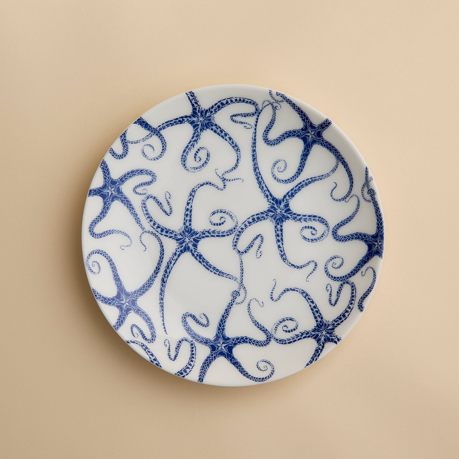 A round white porcelain salad plate featuring a blue starfish motif from Caskata Artisanal Home’s Starfish Coupe Salad Plate collection is displayed against a plain beige background, highlighting its ocean-inspired dinnerware design.