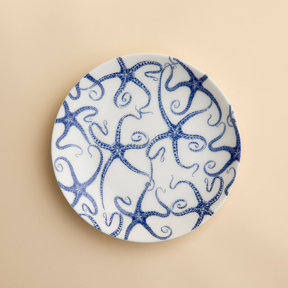 A round white porcelain salad plate featuring a blue starfish motif from Caskata Artisanal Home’s Starfish Coupe Salad Plate collection is displayed against a plain beige background, highlighting its ocean-inspired dinnerware design.