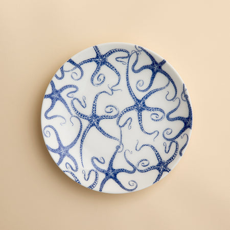 A round white porcelain salad plate featuring a blue starfish motif from Caskata Artisanal Home’s Starfish Coupe Salad Plate collection is displayed against a plain beige background, highlighting its ocean-inspired dinnerware design.