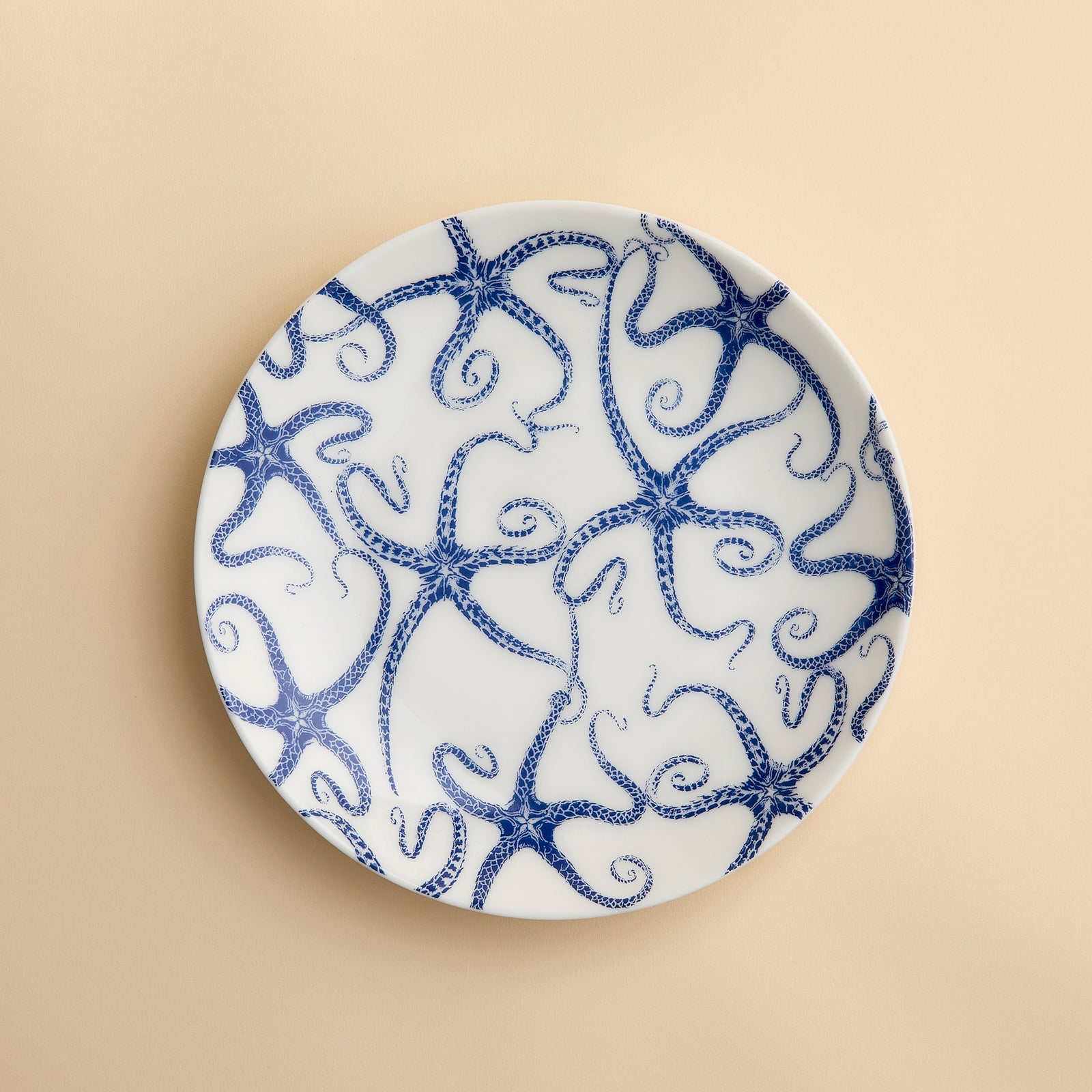 A round white porcelain salad plate featuring a blue starfish motif from Caskata Artisanal Home’s Starfish Coupe Salad Plate collection is displayed against a plain beige background, highlighting its ocean-inspired dinnerware design.